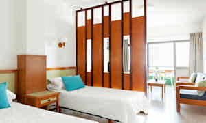 Ett hotellrum med två enkelsängar, ett träbord och en sittgrupp avskild av en rumsdelare. (AI-genererad text)