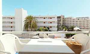 Utsikt från en balkong med sittgrupp, byggnader och palmer synliga i bakgrunden under en klarblå himmel. (AI-genererad text)