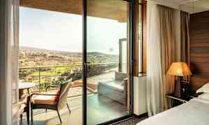 Ett hotellrum med utsikt genom en glasdörr mot en balkong med stolar och ett avlägset landskap utomhus. (AI-genererad text)