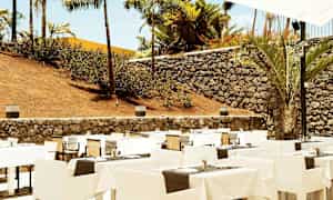 Utomhusrestaurang med uppställda bord och stolar, omgiven av stenmur och palmer. (AI-genererad text)