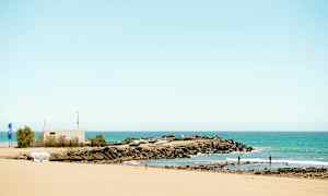 Sandstrand med utsikt över ett klippområde och småbåtshamn vid havet. (AI-genererad text)