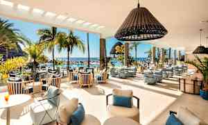 Interiören i en ljus lounge med moderna möbler, stora fönster och en utsikt över en poolområde med palmer och hav. (AI-genererad text)