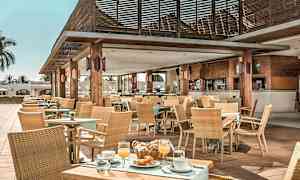 Ett utomhuscaféområde med träbord, stolar och tak, utsmyckat med växter och dukar, serverar mat och dryck. (AI-genererad text)