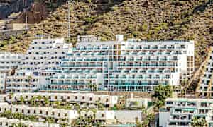 Bild av ett hotell med vit fasad och balkonger, beläget vid en stenig sluttning. (AI-genererad text)