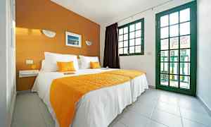 Ett sovrum med en dubbelsäng klädd i vita och orange textilier, och stora fönster med gröna ramar. (AI-genererad text)