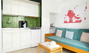 Ett enkelt kök med vit inredning och gröna kakel bakom. Brevid finns en soffa med turkosa dynor och pastellfärgade kuddar. (AI-genererad text)