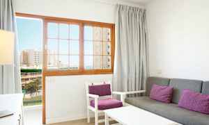 Ett vardagsrum med grå soffa och stolar med lila kuddar, och en trädörr med glaspartier som visar en vy ut mot byggnader och hav. (AI-genererad text)