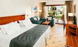 Ett hotellrum med en bred säng, sittgrupp och en uteplats med utsikt över en grönskande trädgård. (AI-genererad text)