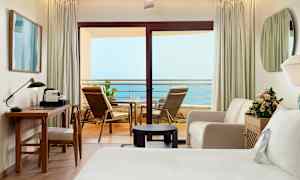 Ett hotellrum med en balkong som har havsutsikt. Rummet är möblerat med skrivbord, stol, soffa, säng och blommor. (AI-genererad text)