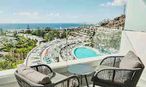 Utsikt från en balkong över ett hotell med en stor pool, omgivande palmer och havet i bakgrunden. (AI-genererad text)