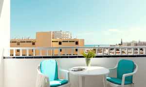 En balkong med två stolar, ett bord och utsikt över byggnader och havet. (AI-genererad text)