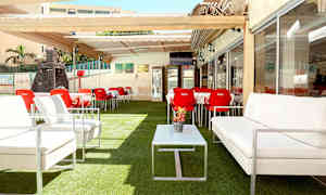 Uteservering med vita soffor, röda stolar och bord, belägen på konstgräs och med pergola. (AI-genererad text)