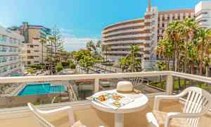 Utsikt över en solig balkong med bord och stolar, hotellkomplex och pool synliga i bakgrunden. (AI-genererad text)