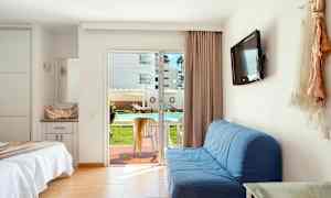 Ett hotellrum med en bäddad säng, blå soffa, väggmonterad TV och en glasdörr som leder ut till en terrass med bord och stolar. Utsikten visar en utomhuspool och solstolar. (AI-genererad text)