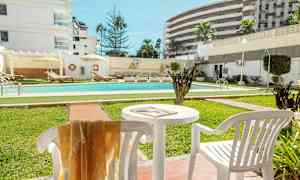 En utomhuspool omgiven av grönytor och solstolar, med vita byggnader i bakgrunden. (AI-genererad text)