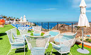 Vita trädgårdsstolar på en gräsyta med havet och landskap i bakgrunden. (AI-genererad text)