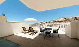 En terrass med solstolar, parasoll och utemöbler, med utsikt mot solen och en bergskulle. (AI-genererad text)