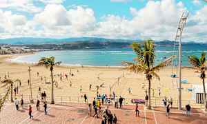En strand med människor som spelar volleyboll och promenerar på strandpromenaden. Havet och bergen syns i bakgrunden. (AI-genererad text)