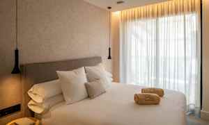 Ett hotellrum med en stor säng, vita lakan och kuddar. På sängen ligger hoprullade handdukar. Väggar och gardiner är ljusa, och rummet har hängande lampor. (AI-genererad text)