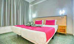 Säng med rosa täcke och kuddar i ett rum med grå gardiner och en träbänk. (AI-genererad text)