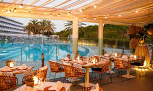 Utomhusrestaurang med dukade bord och ljusslingor, belägen nära en pool och omgiven av palmer och grönska. (AI-genererad text)