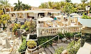 Utomhusrestaurang med vita parasoller och sittplatser, omgiven av grönska och palmer. (AI-genererad text)