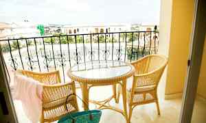 Två rottingstolar och ett runt bord med glasskiva står på en balkong med utsikt över kortstensbyggnader och ett tennisområde. (AI-genererad text)