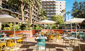 Bilden visar en utomhusrestaurang med färgglada stolar, bord och parasoller framför ett hotell med balkonger. (AI-genererad text)