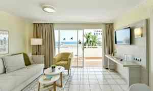 Ett vardagsrum med ljusa möbler och en stor glasdörr som leder till en balkong med utsikt över havet. (AI-genererad text)