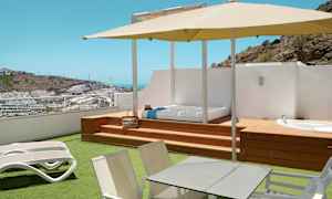 Terrass med solstolar, bord, parasoll samt utsikt över ett kuperat område med byggnader. (AI-genererad text)