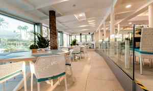 Interiören av en restaurang med vita stolar och bord, glasväggar och utsikt mot grönska utomhus. (AI-genererad text)