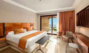 Ett hotellrum med en stor säng, orange inredningsdetaljer, ett skrivbord och en balkong med havsutsikt. (AI-genererad text)