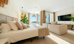Bild av ett ljust och modernt sovrum med stor säng, soffa, TV och en vidsträckt havsutsikt genom stora fönster. (AI-genererad text)