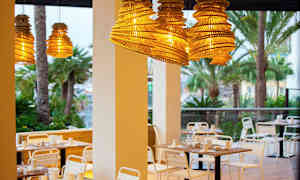 Utemöbler vid restaurang med hängande lampskärmar av trä och palmträd i bakgrunden. (AI-genererad text)