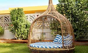 En rottingmöbel i form av en baldakin med randiga blåvita kuddar står på en gräsmatta bredvid några gröna buskar. (AI-genererad text)