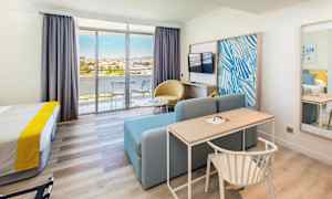 Ett hotellrum med säng, skrivbord, soffa, tv och glasdörrar som leder till en balkong. (AI-genererad text)