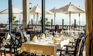 Ett dukat bord i en restaurang med utsikt över en terrass med parasoller och havet. (AI-genererad text)