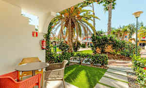 En terrass med bord och färgglada stolar, inramad av en båge som leder ut till en trädgård med palmer och byggnader i bakgrunden. (AI-genererad text)