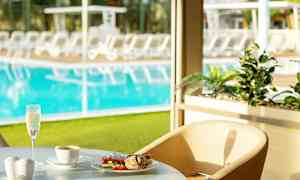 Ett bord nära ett fönster med en tallrik mat, en kopp kaffe och ett glas mousserande dryck. Poolområdet med solstolar syns i bakgrunden. (AI-genererad text)