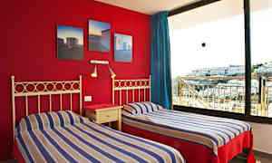 Två enkelsängar med randiga sängkläder och en röd vägg med doft bilder samt ett stort fönster med utsikt över byggnader. (AI-genererad text)