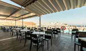 Uteservering med bord och stolar, utsikt över stad och hav. (AI-genererad text)