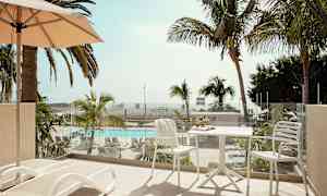 En balkong med solstolar och ett bord med stolar, utsikt över en pool och palmer. (AI-genererad text)