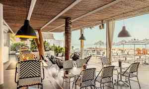 En utomhusservering med rottingmöbler och taklampor, utsikt mot hav och parasoller. (AI-genererad text)