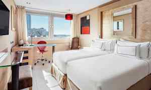 Ett hotellrum med två enkelsängar, ett skrivbord och en stor fönstermiljö som visar stadsutsikt. (AI-genererad text)