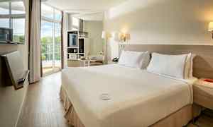 Hotelrum med dubbelsäng och ljus inredning, stora fönster och balkong med utsikt. (AI-genererad text)