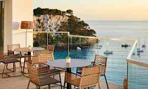 En terrass med bord och stolar, utsikt över havet och båtar vid en klippig kust. (AI-genererad text)