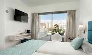 Ett hotellrum med två sängar bäddade med ljusa lakan och turkosa överkast. En stol står framför en glasdörr som leder ut till en balkong. En väggmonterad tv hänger bredvid en hylla med vattenflaskor och kaffemaskin. (AI-genererad text)