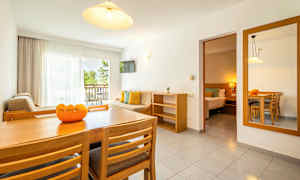 Ett ljust vardagsrum med matbord, stolar, soffa och balkong med utsikt över grönska. Ett anslutande sovrum syns också. (AI-genererad text)