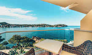 Utsikt från en balkong mot en vik med klart blått vatten, inramad av kuperad terräng och träd. Ett bord och stolar syns i förgrunden på balkongen. (AI-genererad text)