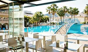 Ett restaurangområde med uteservering vid en pool omgiven av palmer och byggnader. En blå gångbro sträcker sig över poolen. (AI-genererad text)